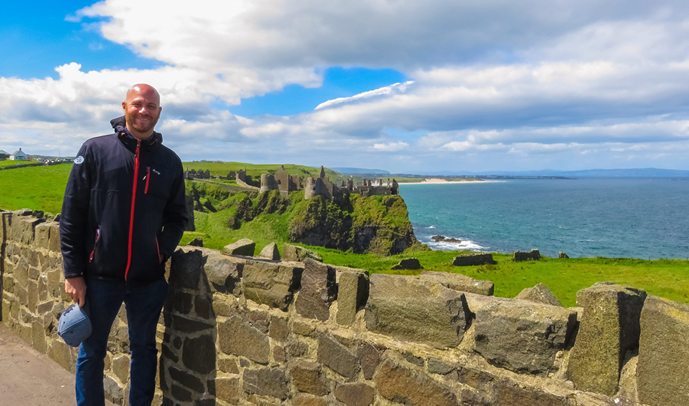 On a sunny day I'm standing in front of a low brick wall overlooking the blue Northern Irish sea and ruins of a gray Medieval castle that was part of the Game of Thrones television show that sits on lush green land
